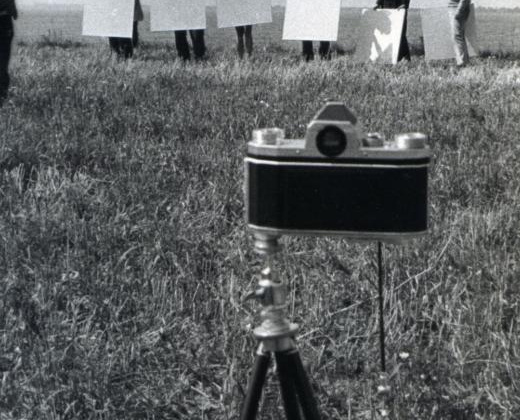 zdjęcie czarno białe, zdjęcie zrobione na dworzu, na pierwszym planie aparat fotograficzny starego typu, w tle 6 osób