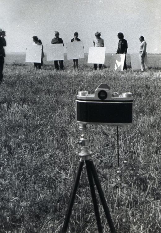zdjęcie czarno białe, zdjęcie zrobione na dworzu, na pierwszym planie aparat fotograficzny starego typu, w tle 6 osób